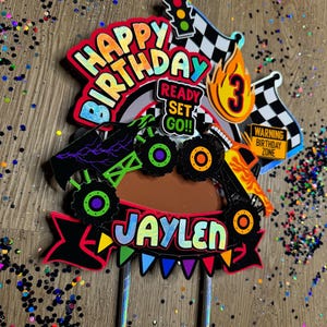 May include: A colorful monster truck cake topper with the words "Happy Birthday" and "Jaylen" in rainbow letters. The topper also includes a checkered flag, a traffic light, a flame with the number 3, and a sign that says "Warning Birthday Zone".