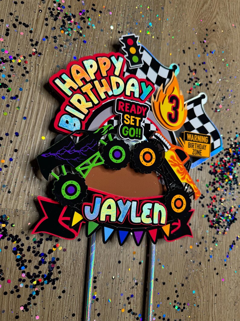 May include: A colorful monster truck cake topper with the words "Happy Birthday" and "Jaylen" in rainbow letters. The topper also includes a checkered flag, a traffic light, a flame with the number 3, and a sign that says "Warning Birthday Zone".
