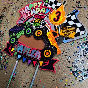 May include: A monster truck cake topper with a red, yellow, and black monster truck, a red and yellow banner that says "Happy Birthday", a red, yellow, and blue banner that says "Ready Set Go!!", a yellow and orange banner that says "Jaylen", a yellow and orange number 3, a red and white checkered flag, a red and white checkered flag with a yellow flame, and a yellow sign that says "Warning Birthday Zone".