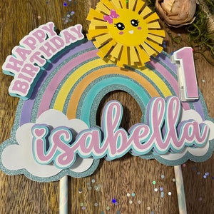 Może przedstawiać: Topper na tort w kształcie tęczy z słońcem, chmurami i napisami "Happy Birthday" i "Isabella 1".