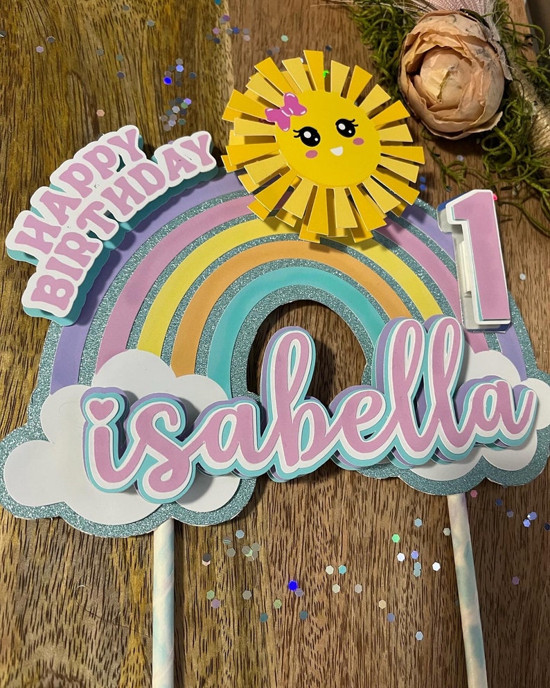 Może przedstawiać: Topper na tort w kształcie tęczy z słońcem, chmurami i napisami "Happy Birthday" i "Isabella 1".
