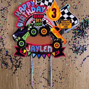May include: A colorful monster truck themed cake topper with the words "Happy Birthday", "Ready Set Go!!", and "Jaylen". The topper also features a checkered flag, a traffic light, a number 3, and a monster truck.