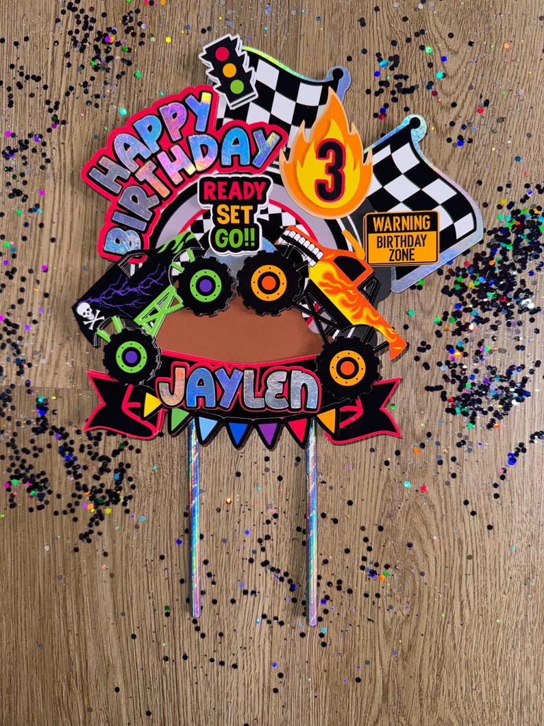 May include: A colorful monster truck themed cake topper with the words "Happy Birthday", "Ready Set Go!!", and "Jaylen". The topper also features a checkered flag, a traffic light, a number 3, and a monster truck.