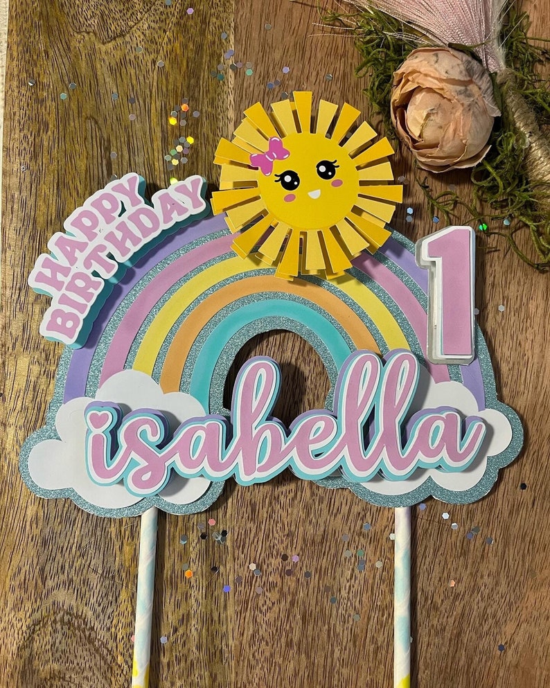 Może przedstawiać: Topper na tort w kształcie tęczy z słońcem, chmurami i napisami "Happy Birthday" i "Isabella" w kolorze r&oacute;żowym. Liczba "1" r&oacute;wnież jest w kolorze r&oacute;żowym.