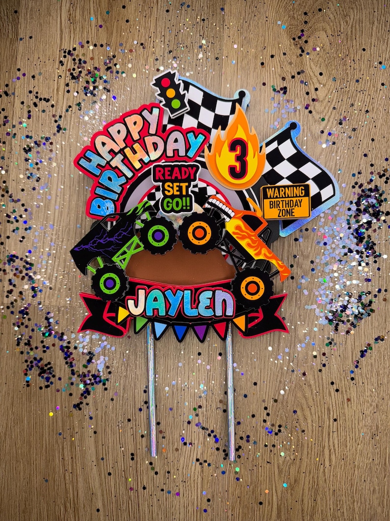 May include: A colorful monster truck cake topper with the words "Happy Birthday" and "Ready Set Go!!" in a rainbow font. The topper also includes a checkered flag, a traffic light, a number 3, and the name "Jaylen".