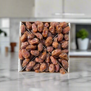 May include: A square cardboard box filled with numerous brown dates. The dates are a rich, caramel color and have a wrinkled texture. The box is divided into sections, and the background is a blurred kitchen setting.