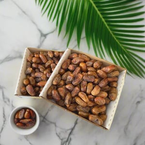 May include: Two cardboard boxes filled with brown dates, alongside a small white bowl of dates. A green palm leaf is in the background. The dates are a natural, healthy snack.