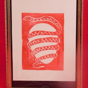 May include: A framed print of a red and white linocut print of a snake coiled around an egg.