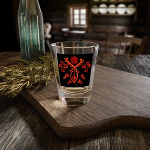 Può includere: Un bicchierino di vetro trasparente con un designo nero e rosso di una testa di cervo stilizzata con corna e forme geometriche. Il bicchierino è appoggiato su una superficie di legno.