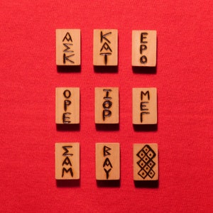 Puede incluir: Nueve fichas de madera con diferentes símbolos y letras escritas en ellas. Las fichas están dispuestas en una cuadrícula de 3x3 sobre una superficie roja. Los símbolos incluyen las letras "A", "E", "I", "K", "M", "O", "P", "S", "T" e "Y".