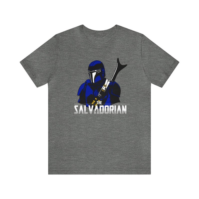 May include: Gray t-shirt featuring a blue and black illustration of a helmeted figure with a weapon. The text "THE SALVADORIAN" is printed in white below the figure. The shirt is made of a soft material.