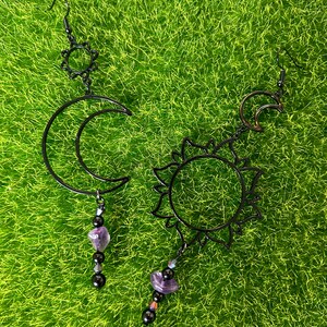 May include: A pair of black metal earrings with a crescent moon and sun design. Each earring features a dangling amethyst stone and black beads.
