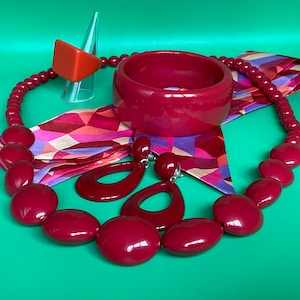 May include: A set of red plastic jewellery including a necklace, bracelet, and earrings. The necklace and bracelet are made of large, oval-shaped beads. The earrings are large and dangle.