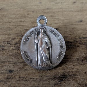 May include: A silver Saint Maurus medal with a loop for hanging. The medal features an image of a robed saint holding a staff and book, with the words "SAINT MAUR" and "PRIEZ POUR NOUS" around the edge.