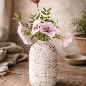 May include: A textured, off-white vase holds a bouquet of pink and purple petunias and greenery. A tall, reddish-brown grass-like stem rises above the flowers. The vase sits on a rustic wooden surface, suggesting a natural, home decor theme.