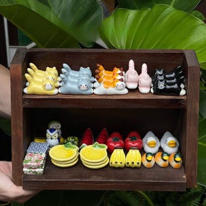 Puede incluir: Una estantería de madera exhibe una colección de figuritas de cerámica. La estantería superior presenta figuritas de gatos en amarillo, azul, naranja, rosa y negro. La estantería inferior contiene figuritas de búhos, frutas y patos.