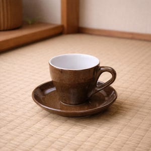 Può includere: Un set di tazza e piattino in ceramica marrone. La tazza ha un interno bianco e un manico curvo. Il piattino è rotondo e si abbina al colore della tazza. Il set è posizionato su una superficie chiara e strutturata.