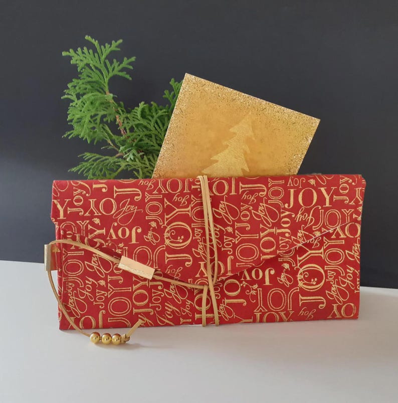 Op de afbeelding: Een rode geschenkdoos met gouden "JOY" tekst, vastgebonden met een lichtbruin koord en een klein houten accent. Een gouden glitterkaart met een kerstboomontwerp is ingevoegd en een takje groen loof voegt een natuurlijke toets toe.