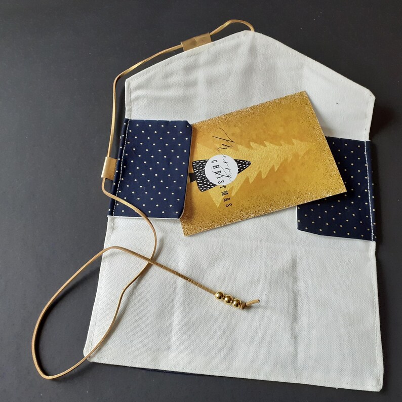 Op de afbeelding: Witte stoffen clutch met een gouden koordriem en gouden kraalaccenten. De binnenkant heeft marineblauwe polka dot zakken en een gouden kerstkaart met een boomontwerp en de tekst "Merry Christmas".