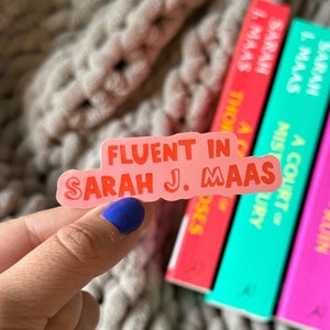 May include: A pink sticker with the text "Fluent in Sarah J. Maas" in red lettering. The sticker is being held by a hand with blue nail polish. The sticker is in front of a stack of books with the author's name Sarah J. Maas on the spines.