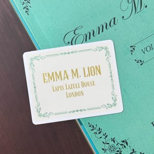 May include: A white rectangular card with gold text that reads "EMMA M. LION LAPIS LAZULI HOUSE LONDON" is placed on a teal book. The book has the title "Emma M. Lion" in elegant script and "VOL. 1" in a box.