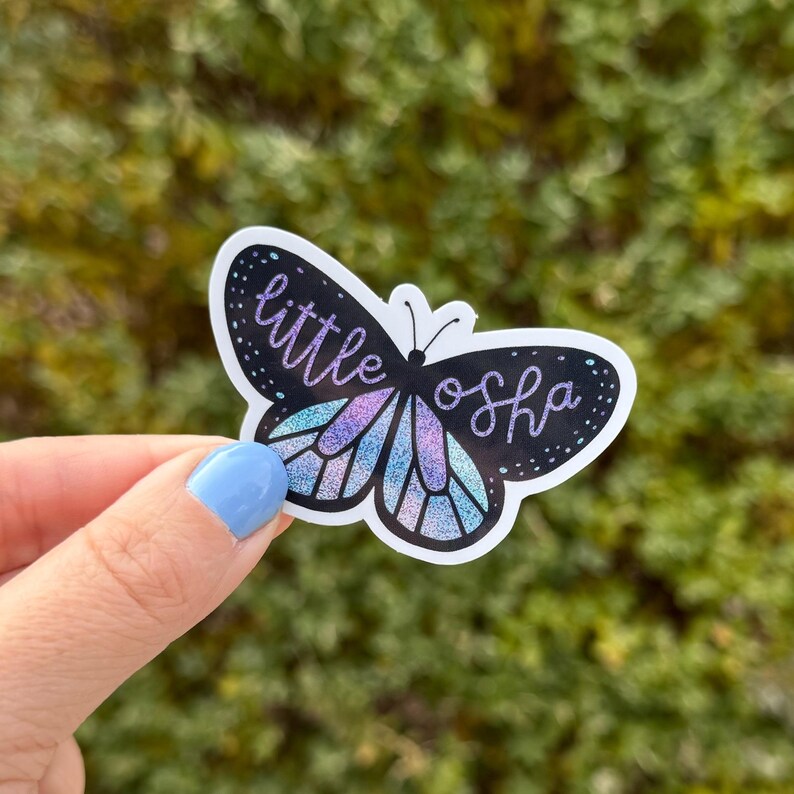 May include: A sticker featuring a purple and blue butterfly with glitter accents. The butterfly has black wings with white outlines and the text "little osh" written on the wings.