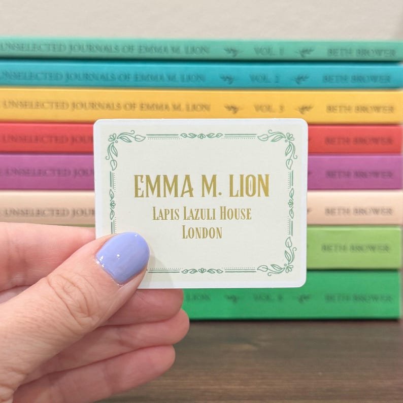 May include: A cream-colored rectangular sticker with the text "EMMA M. LION, LAPIS LAZULI HOUSE, LONDON" in gold lettering, framed by a green floral border. A stack of colorful books is in the background.