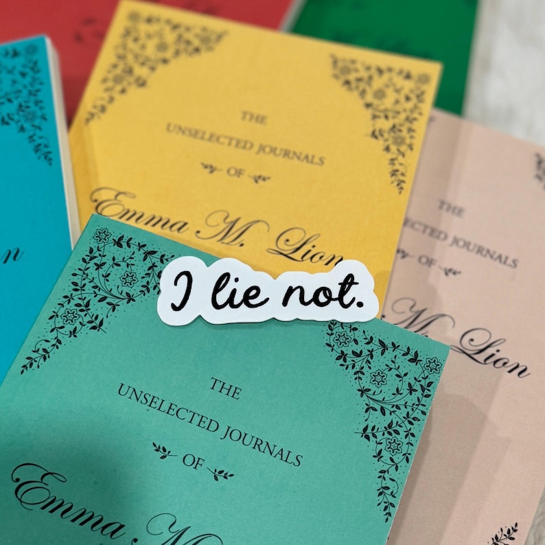 Op de afbeelding: Een verzameling tijdschriften in verschillende kleuren, waaronder teal, geel en perzik. Elk tijdschrift bevat de tekst "The Unselected Journals of Emma M. Lion" en decoratieve bloemenhoekontwerpen. Een witte sticker met "I lie not." is erop geplakt.