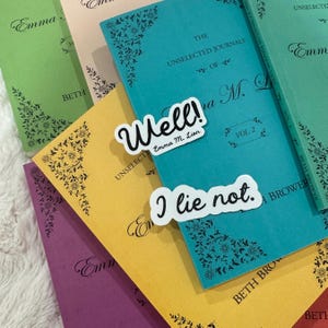 Op de afbeelding: Een verzameling kleurrijke boeken met bloemenhoekontwerpen en de titel "The Unselected Journals of Emma M. Lion". Twee stickers lezen "Well!" en "I lie not." De boeken zijn in tinten van teal, geel, paars, groen en roze.