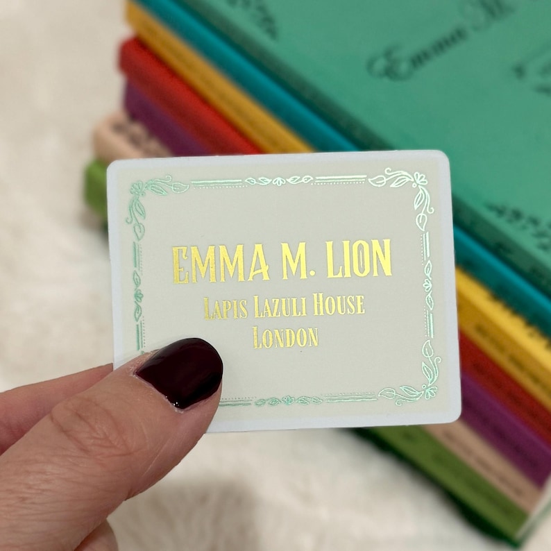 May include: A rectangular business card with the text "EMMA M. LION, LAPIS LAZULI HOUSE, LONDON" in gold lettering. The card has a white background with a decorative green border. A stack of colorful books is in the background.
