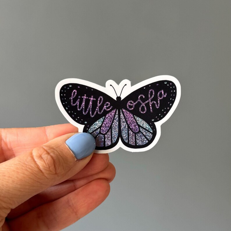 May include: A black butterfly sticker with iridescent wings and the text "little osha" in pink glitter.