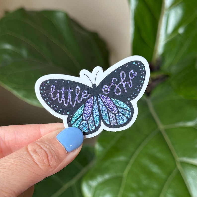 May include: A sticker of a butterfly with iridescent wings and the text "little osh" in black script.