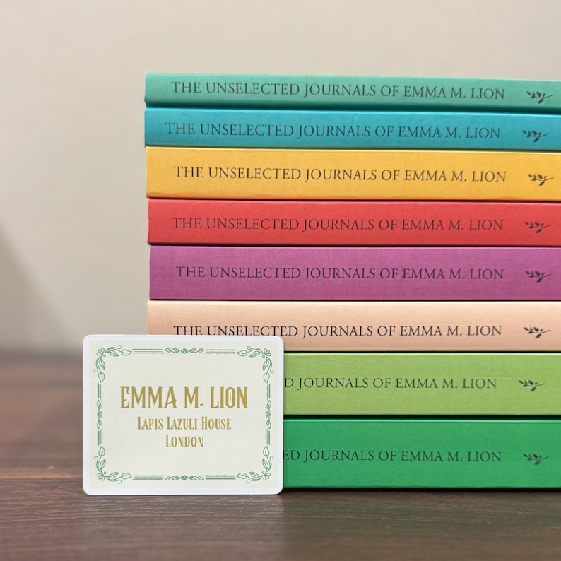 May include: A stack of colorful journals titled "The Unselected Journals of Emma M. Lion" with a decorative card in front. The card reads "Emma M. Lion, Lapis Lazuli House, London."