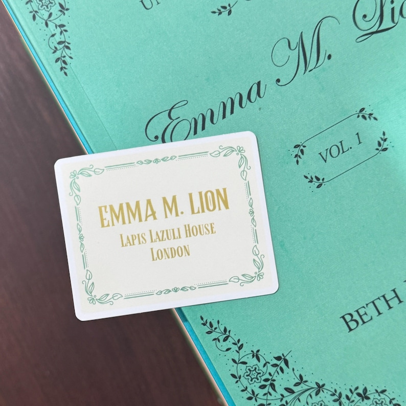 May include: A white rectangular card with gold text that reads "EMMA M. LION LAPIS LAZULI HOUSE LONDON" is placed on a teal book. The book has the title "Emma M. Lion" in elegant script and "VOL. 1" in a box.