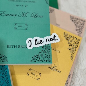 Op de afbeelding: Een stapel tijdschriften in teal, geel en perzik. Het teal tijdschrift heeft de tekst "Emma M. Lion" en "Vol. 1". Een witte sticker met de tekst "I lie not." ligt erop. Bloemenhoekontwerpen zijn zichtbaar.