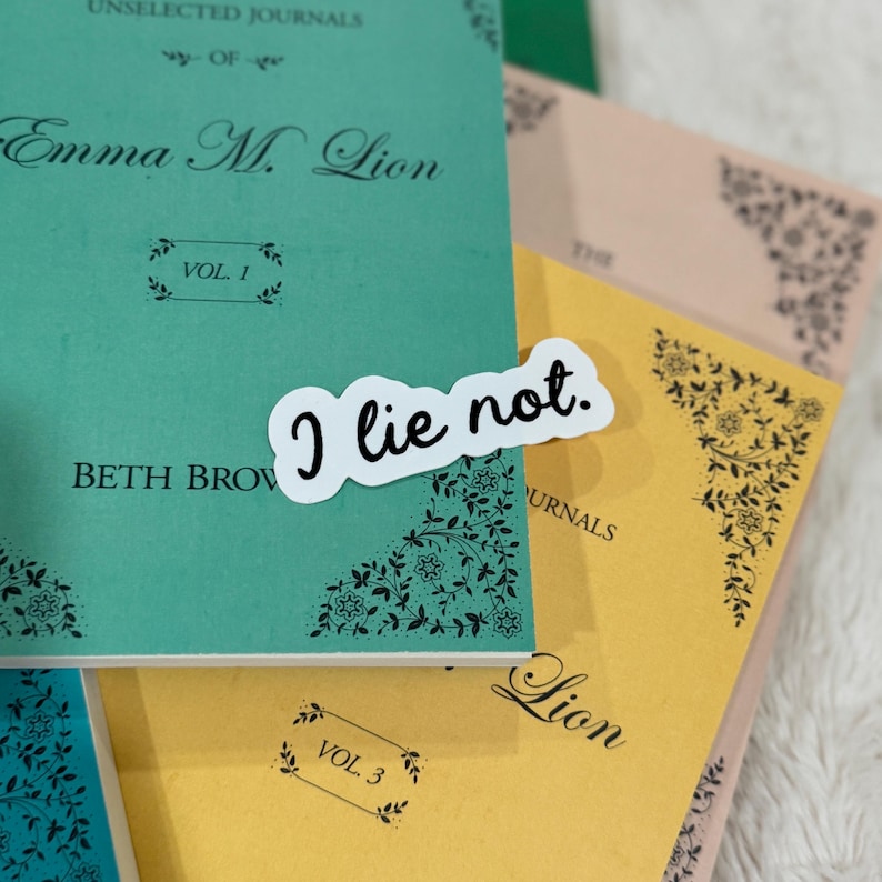 Op de afbeelding: Een stapel tijdschriften in teal, geel en perzik. Het teal tijdschrift heeft de tekst "Emma M. Lion" en "Vol. 1". Een witte sticker met de tekst "I lie not." ligt erop. Bloemenhoekontwerpen zijn zichtbaar.