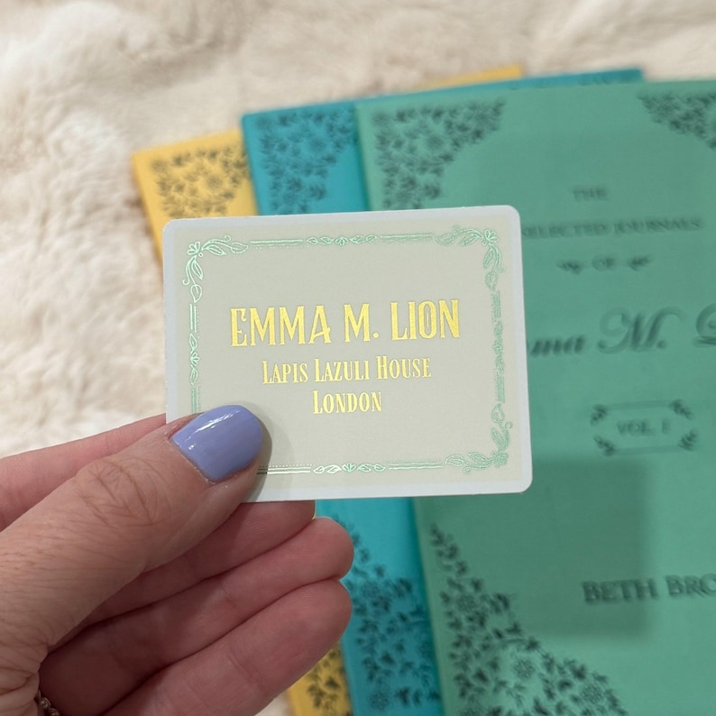 May include: A rectangular sticker with the text "EMMA M. LION, LAPIS LAZULI HOUSE, LONDON" in gold lettering. The sticker has a light green border and is held in front of a stack of books with teal and yellow covers.