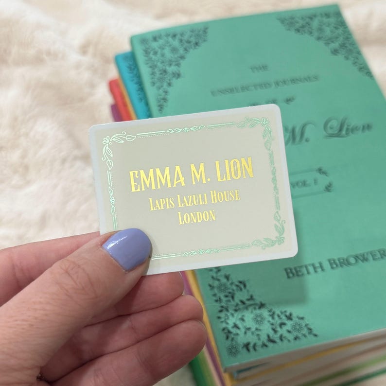 May include: A business card with the name "EMMA M. LION" in gold lettering, along with "LAPIS LAZULI HOUSE LONDON". The card is held in front of a stack of teal books with gold lettering and decorative borders.