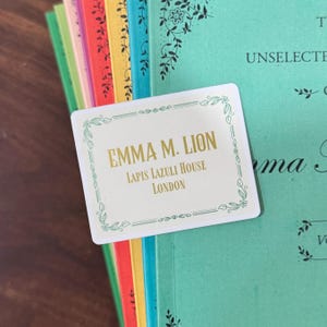 May include: A white card with gold text that reads "EMMA M. LION, LAPIS LAZULI HOUSE, LONDON" is placed on top of a stack of colorful books. The card has a decorative border with floral elements. The books have various colored spines.