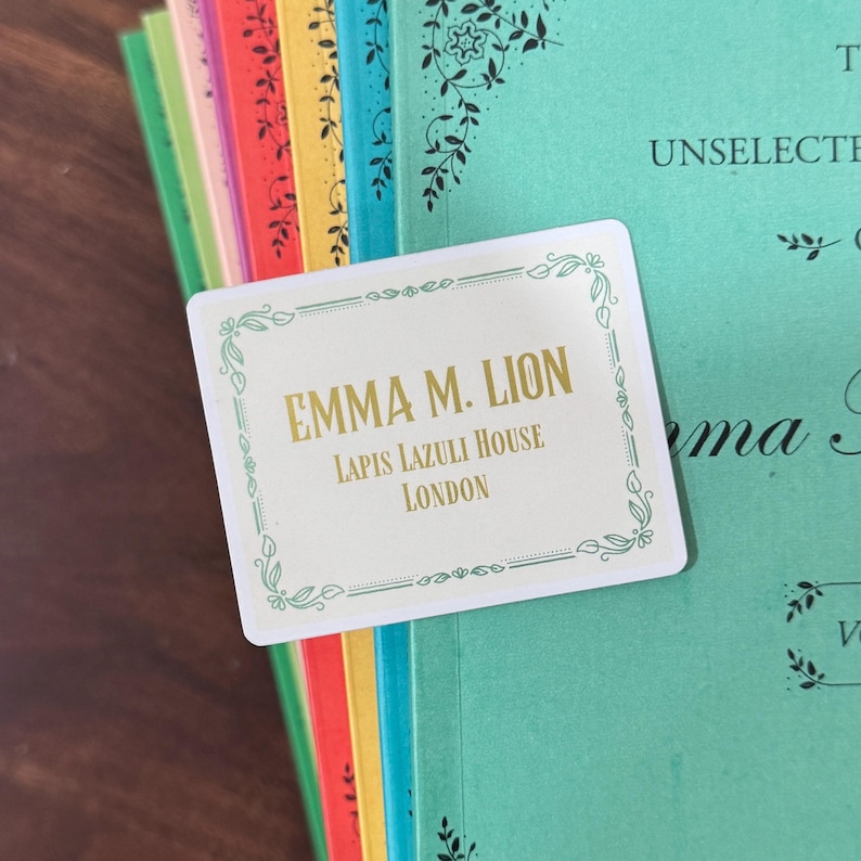 May include: A white card with gold text that reads "EMMA M. LION, LAPIS LAZULI HOUSE, LONDON" is placed on top of a stack of colorful books. The card has a decorative border with floral elements. The books have various colored spines.
