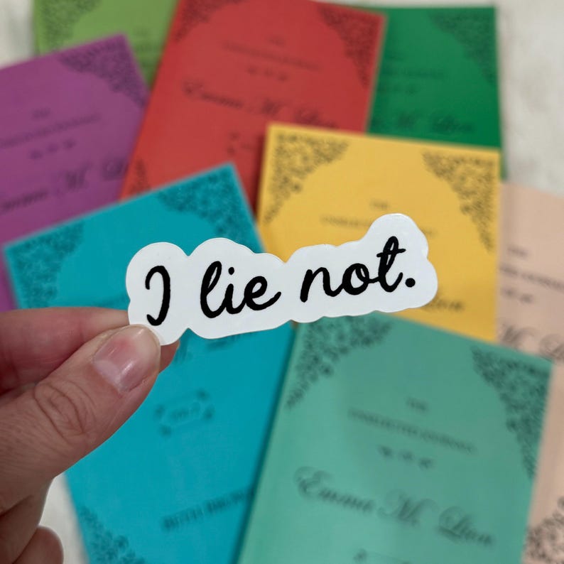 Op de afbeelding: Een witte sticker met de tekst "I lie not." in een zwart, handgeschreven lettertype. De sticker wordt voor een verzameling kleurrijke boeken gehouden, waaronder paars, rood, groen, geel en turquoise.