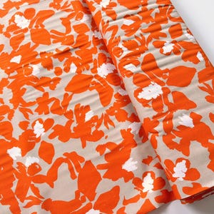 Könnte beinhalten: Eine Stoffrolle mit einem floralen Muster in Orange und Weiß auf einem neutralen beigefarbenen Hintergrund. Das Design zeigt große, stilisierte Blumen und Blätter und erzeugt einen kräftigen und lebendigen Stoff.