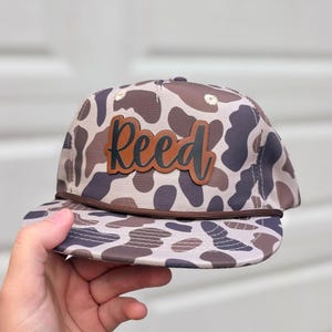 May include: A baseball cap featuring a brown and tan camouflage pattern with a brown cord running across the base of the crown. A rectangular brown leather patch is centered on the front, displaying the name Reed in black cursive lettering.