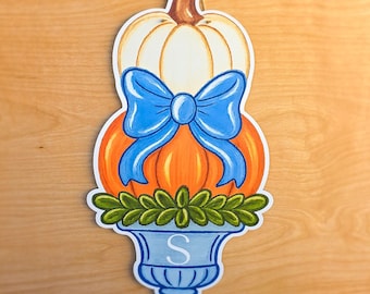 May include: A hanging wall decoration featuring a white pumpkin stacked on an orange pumpkin, accented with a bright blue bow. Below the pumpkins is a green leafy arrangement inside a light blue urn-shaped base with the letter S printed on it.