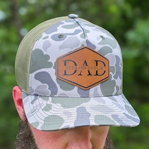 May include: A camouflage print baseball cap with a brown leather patch that reads "DAD" and "REED + MAGNOLIA". The cap has a green mesh back and a curved brim. The hat is a trucker style.