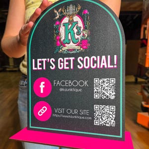 May include: An arched black sign with a teal border and a bright pink base. The sign features the text LET'S GET SOCIAL!, a K's Junktique logo with a deer, and two QR codes with icons for Facebook and a website link for ksjunktique.com.