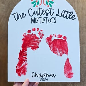 Pode incluir: Placa branca arqueada com pegadas vermelhas e o texto "The Cutest Little Mistletoes" e "Christmas 2024". Um design festivo de la&ccedil;o e bagas no topo. Uma m&atilde;o segura a placa.