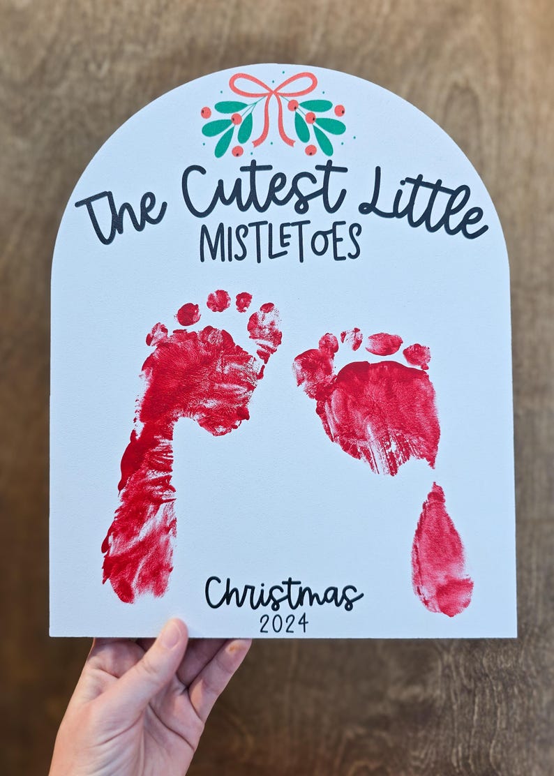 Pode incluir: Placa branca arqueada com pegadas vermelhas e o texto "The Cutest Little Mistletoes" e "Christmas 2024". Um design festivo de la&ccedil;o e bagas no topo. Uma m&atilde;o segura a placa.