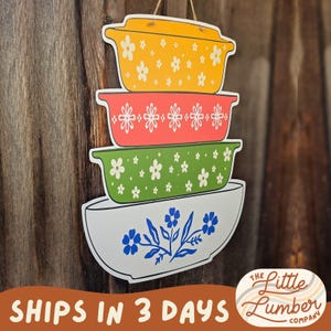 May include: A wooden sign featuring a stack of colorful, retro-style casserole dishes. The top dish is yellow with white floral accents, followed by red, green, and a white bowl with blue floral designs. The sign hangs from a string.