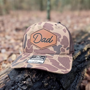 May include: A camouflage trucker hat with a brown mesh back and a leather patch that says "Dad". The hat has a curved brim and a tag that reads "Richardson 112 Trucker".